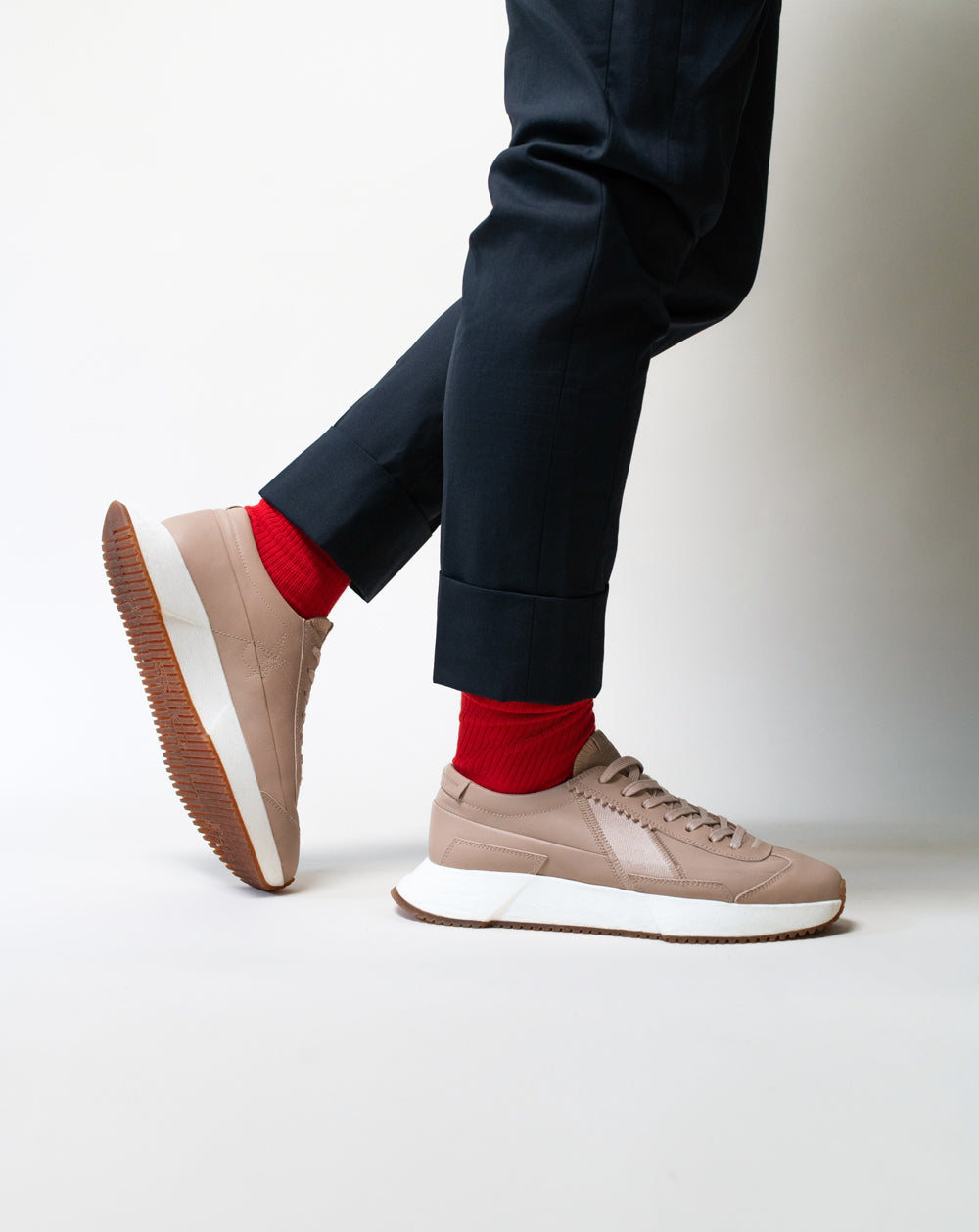 Person wearing beige sneakers with white soles, dark pants, and red socks on a light background