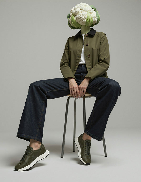 Person wearing a green jacket and dark pants sitting on a stool with a cauliflower head.