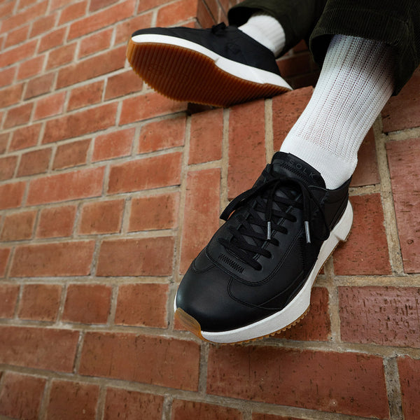 Black sneakers with white soles worn against a brick wall.