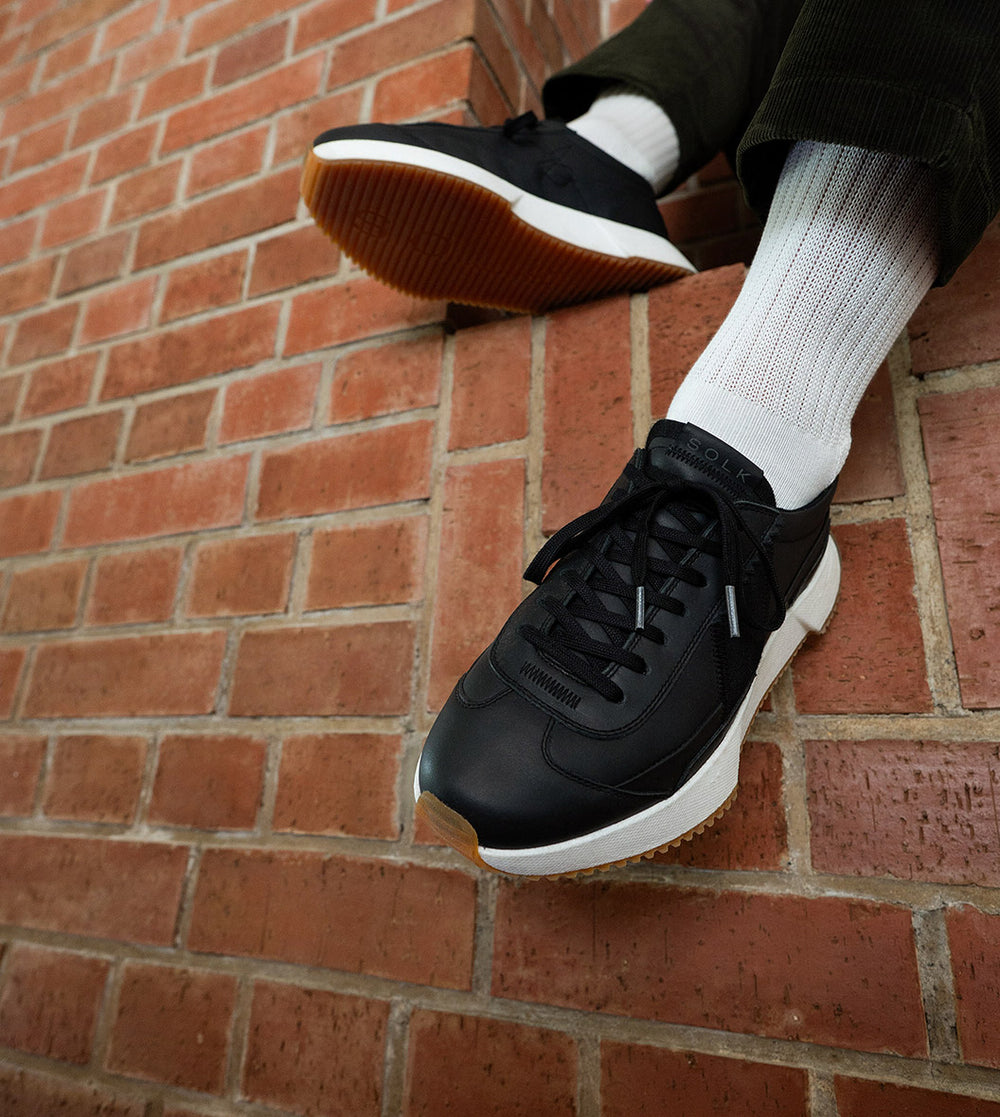 Black sneakers with white soles worn against a brick wall.