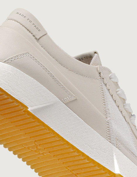 Close-up of a beige sneaker with a white sole and yellow accent on a light gray background.