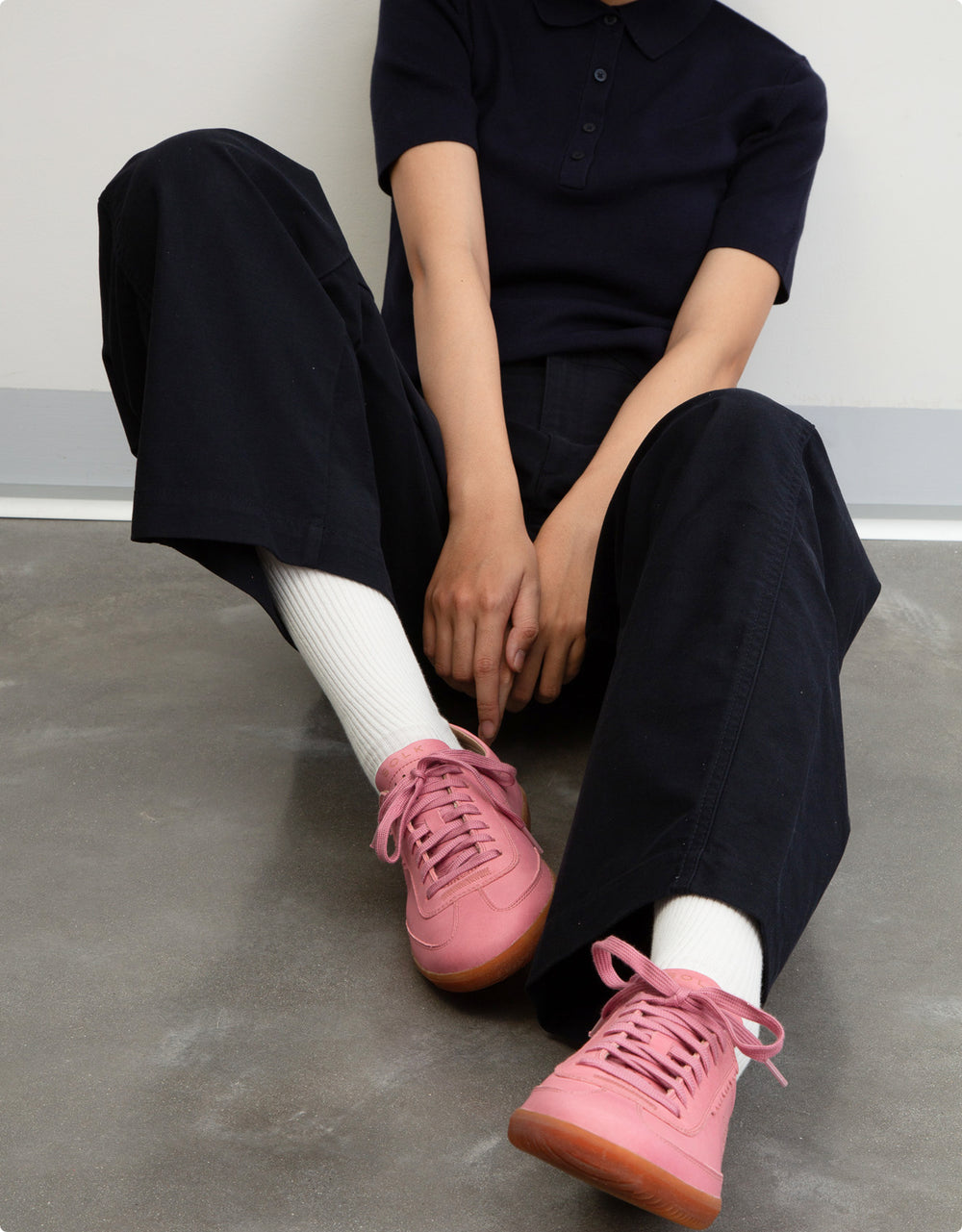 Person wearing a black outfit with pink sneakers sitting on a concrete floor.